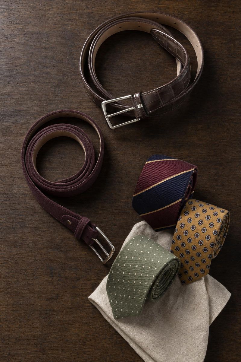 Alligator and suede belts coiled beside rolled silk neckties and a linen pocket square
